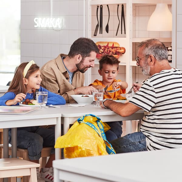 Two adults and two kids talking and laughing while having a meal in an IKEA Restaurant next to a yellow FRAKTA bag.