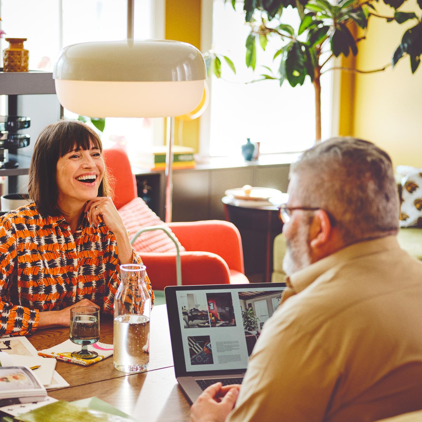 Two adults sit across a wooden table. One is laughing and the other is on a laptop. A glass and water jug sit between them.
