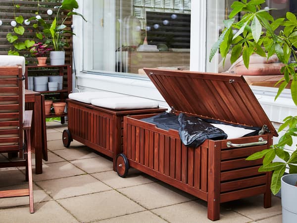 Two ÄPPLARÖ / TOSTERÖ benches in acacia in front of two windows. One bench lid is open and a storage bag is stored inside.