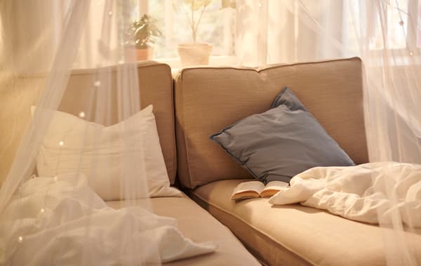 Two beige, side-by-side KIVIK chaise lounges by a sunlit window. Bedspreads and a book on them, a SOLIG net draped over.