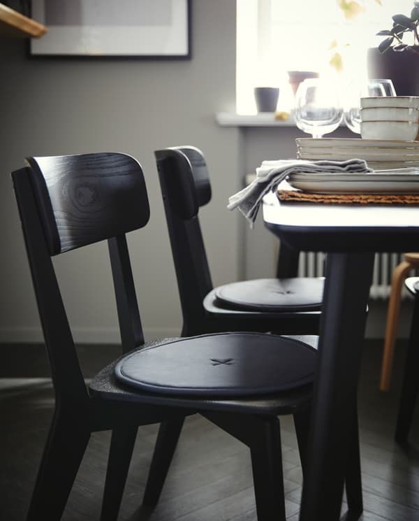 Two black chairs with round, dark grey chair pads stand by a black table where wine glasses and grey dinnerware stand.
