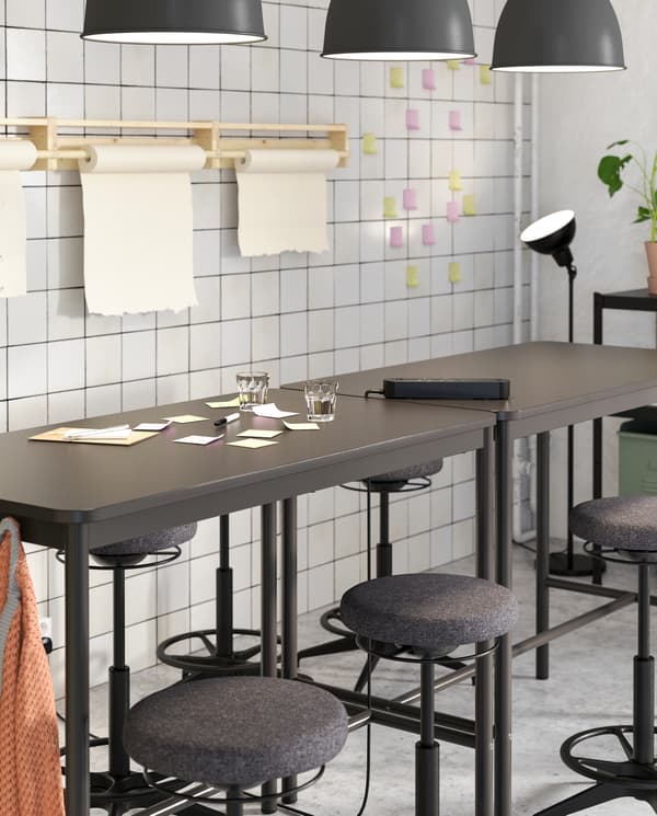 Two black high tables and several black high stools standing together, shown next to a white wall and colourful sticky notes.
