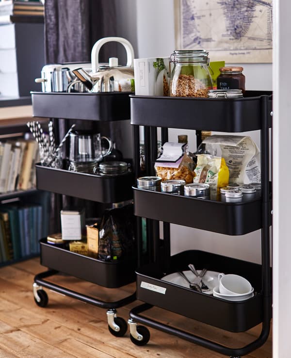 Two black RÅSKOG trolleys full of kitchen items, such as food, a toaster, a kettle and plates.