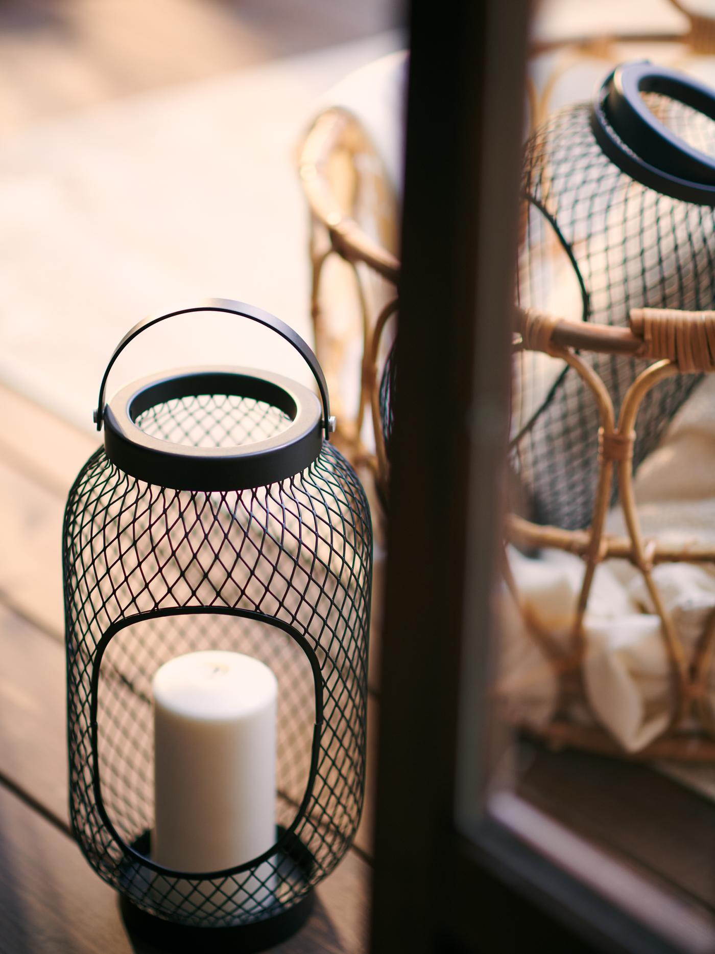 Two black TOPPIG lanterns for block candles placed on a wooden outdoor space together with a SNIDAD rattan basket.