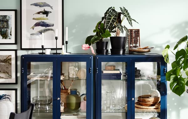 Two blue FABRIKÖR glass-door display cabinets displaying mementos, with plants and candles on top.