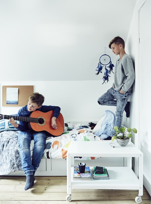 Two boys playing in a child’s bedroom, one standing on the bed, the other playing a guitar.