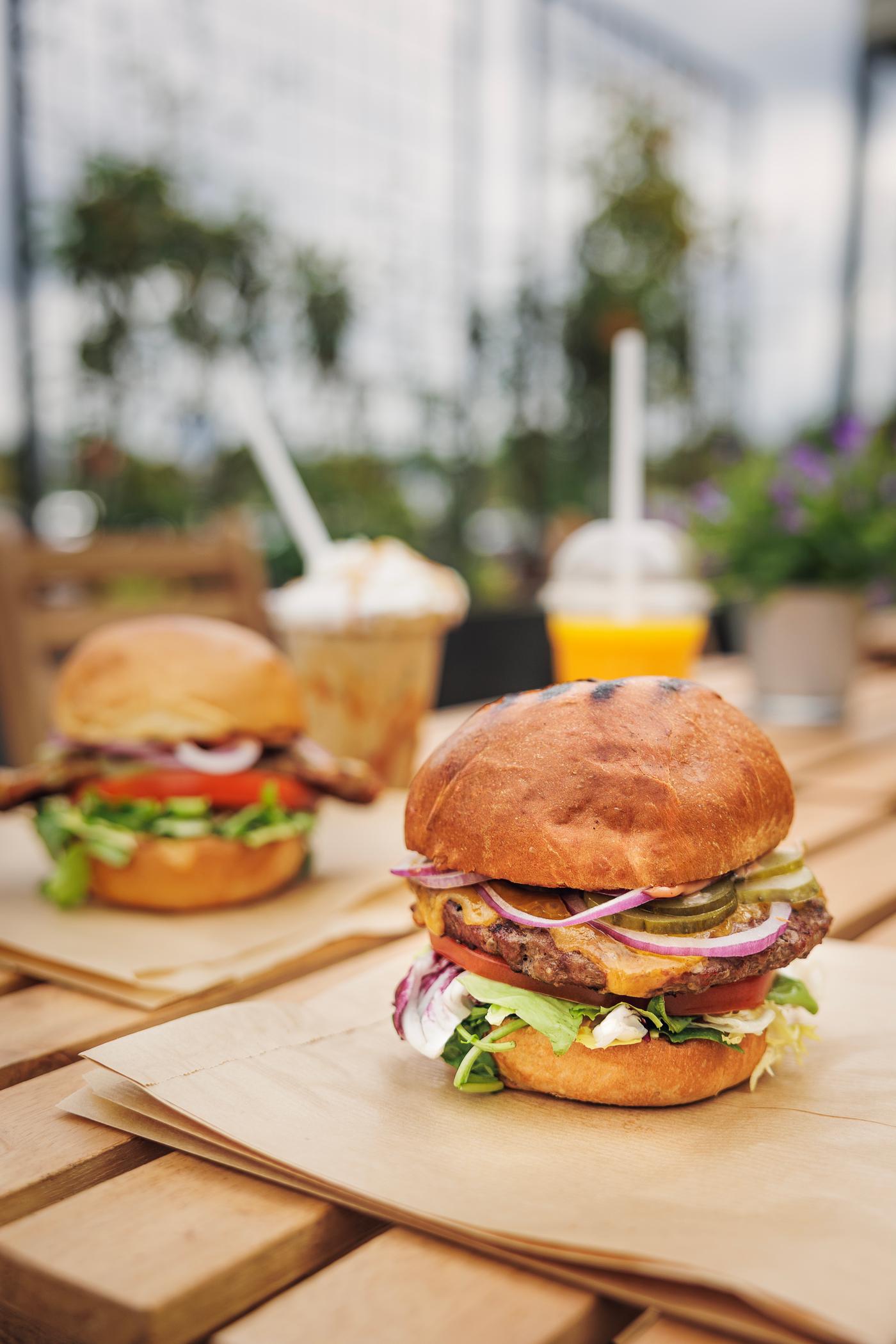Two burgers on a wooden table outdoors, served with iced coffee and peach-mango smoothie.