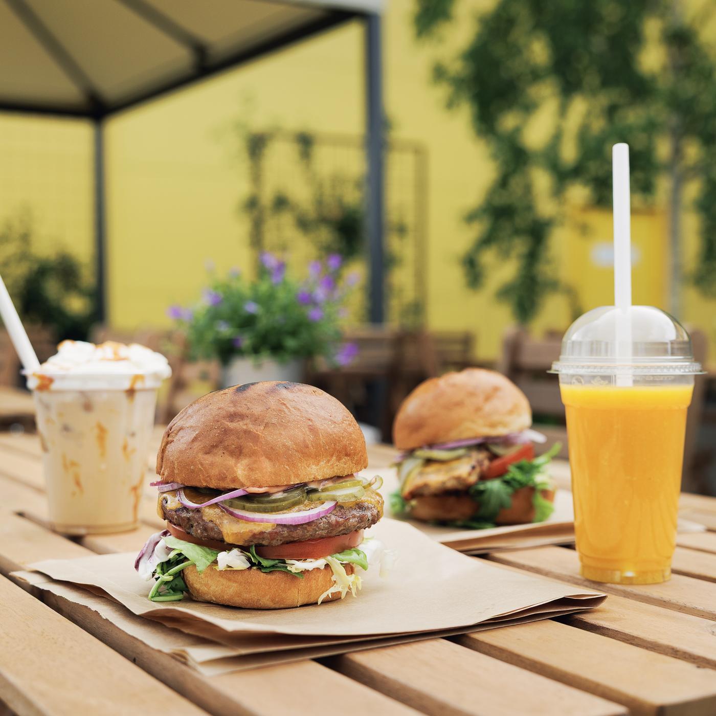 Two burgers on a wooden table outdoors, served with iced coffee and peach-mango smoothie.