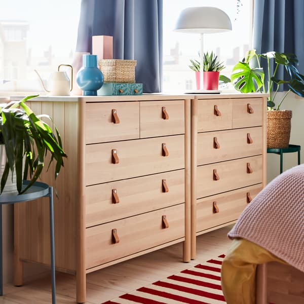 Two chests of drawers in birch with tanned leather handles, a tray table in blue, a white table lamp and some green plants.