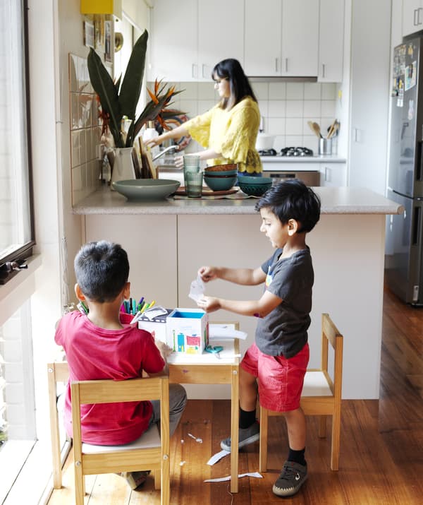 Two children doing crafts at a small table in front of an L-shaped kitchen, where Abeer stands at the sink.