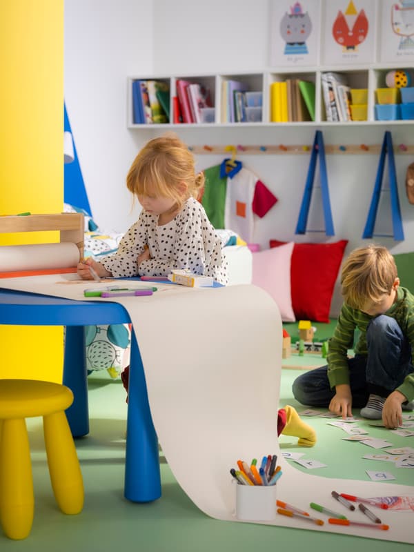 Two children in a bright, modern play area; one sits at a table drawing while the other kneels on the floor among scattered crayons and toys.