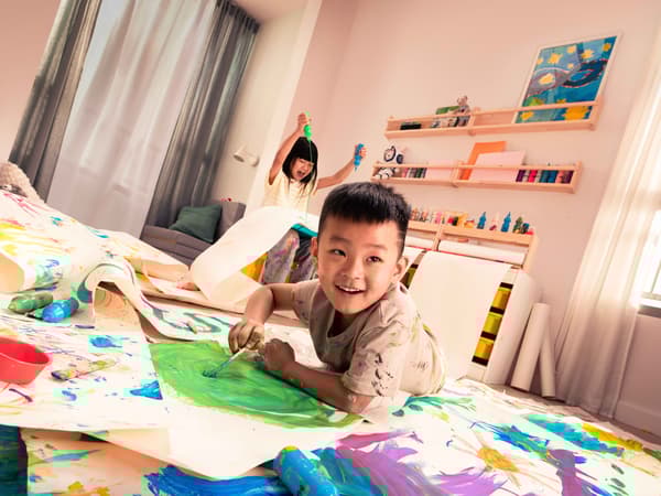 Two children playing in a room, painting with various colours on paper rolls on the floor