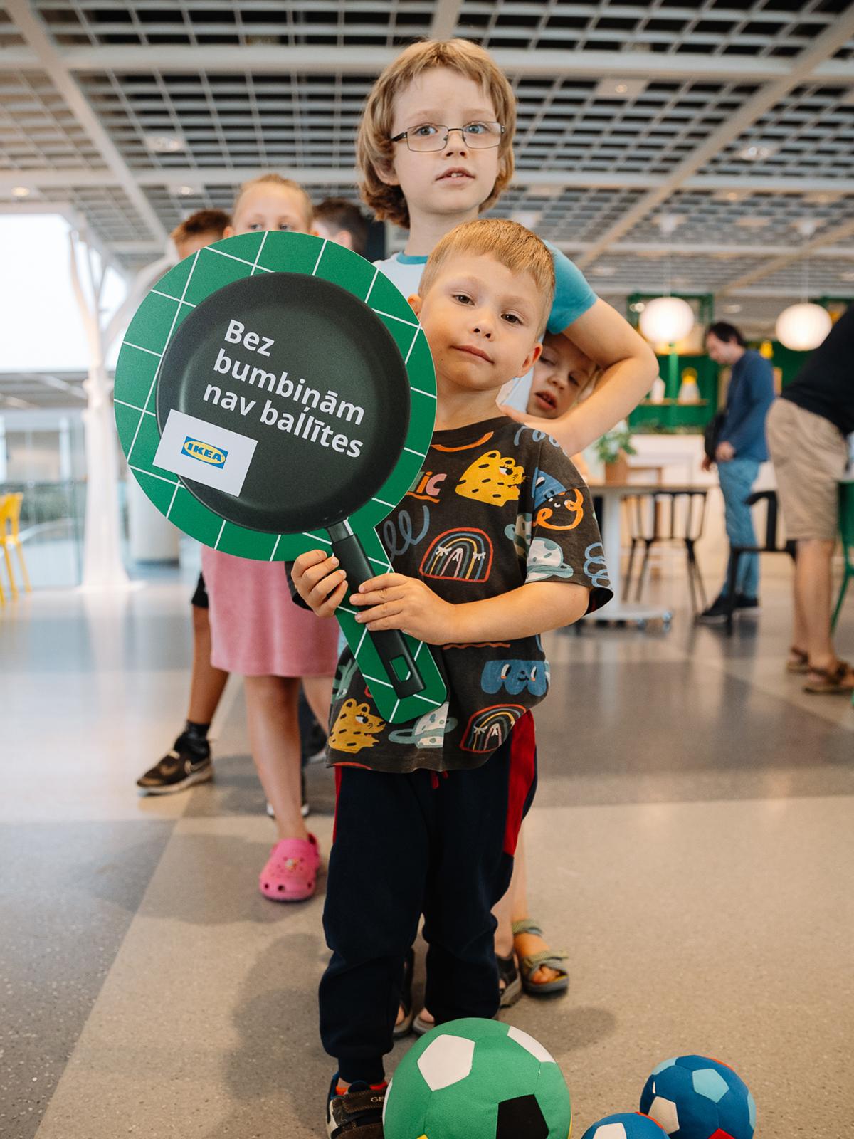 Two children pose with a round event sign beside colourful balls in an IKEA store.