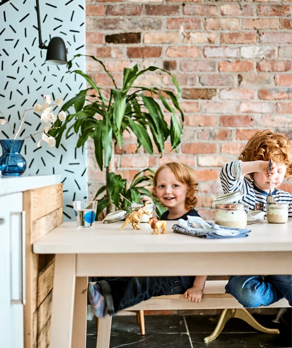Two children sit at a light wooden table with breakfast and toys, in front of a red brick wall.
