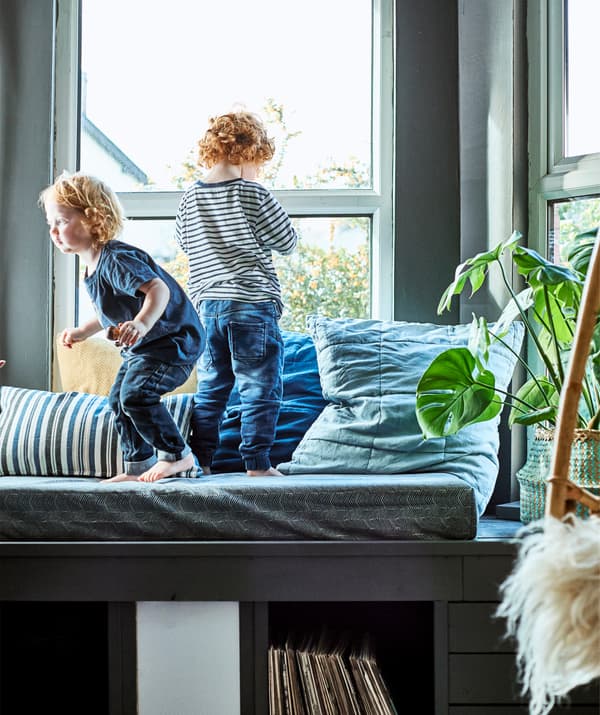 Two children standing on a window seat with large cushions and storage underneath.