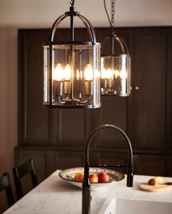 Two cylinder-shaped black/glass pendant lamps hang over a white marble effect worktop, a black mixer tap and a white sink.