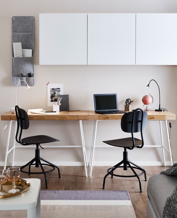 Two desks with light wood worktops in a living room area, sitting against a wall and beneath white wall cabinets.