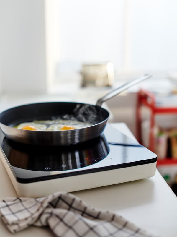 Two eggs frying in a pan on a TILLREDA portable induction hob set on a worktop, a kitchen towel lying beside it.