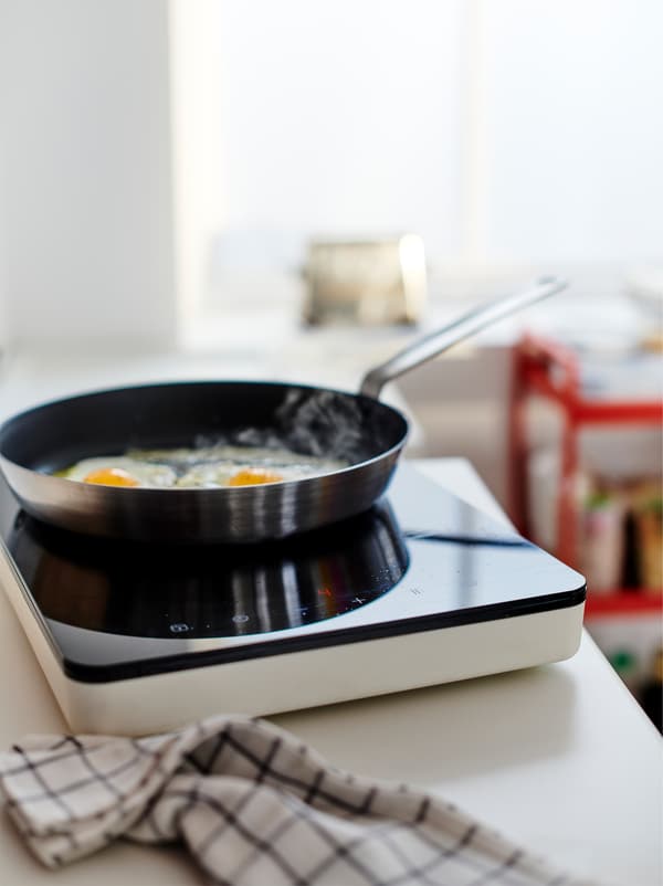Two eggs frying in a pan on a TILLREDA portable induction hob set on a worktop, a kitchen towel lying beside it.