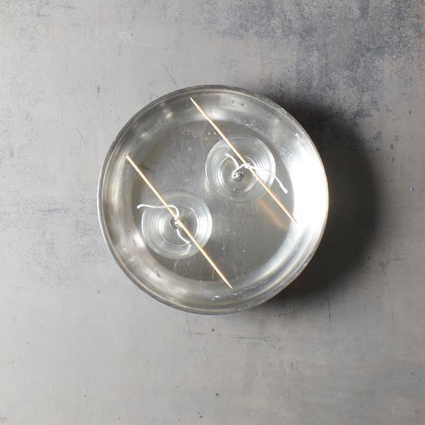 Two glass holders with wicks attached float in warm water, supported by wooden sticks, as if an IKEA craft project were in the middle of the candle-making process.