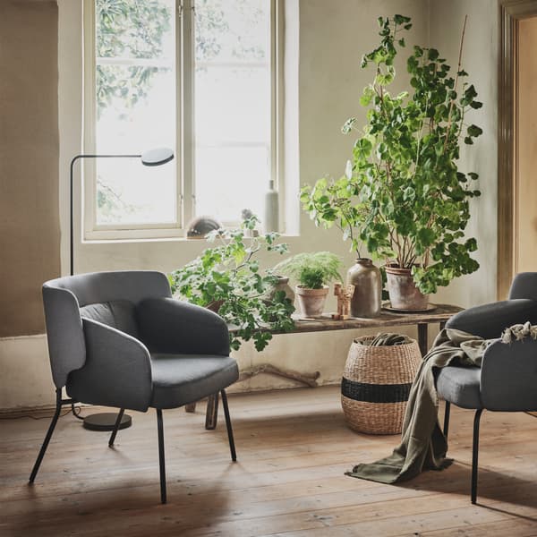 Two gray low back BINGSTA armchairs are seen in a cosy, sunny corner beside a window and a bench filled with plants.