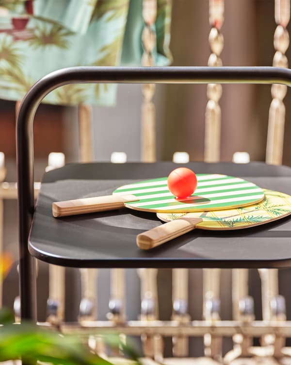 Two green and yellow printed rackets and a ball lying on a black table, standing on a balcony.