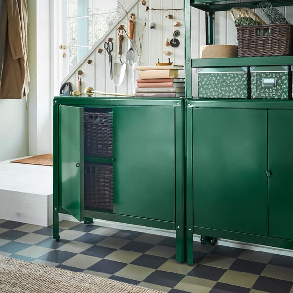 Two green KOLBJÖRN cabinets stand in front of a staircase. One cabinet door is open and inside are dark brown rattan baskets.