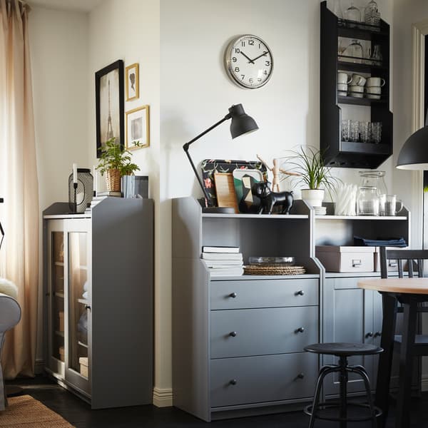 Two grey chests of drawers with shelves with decorative items, a round table, a black chair and a black pendant lamp.