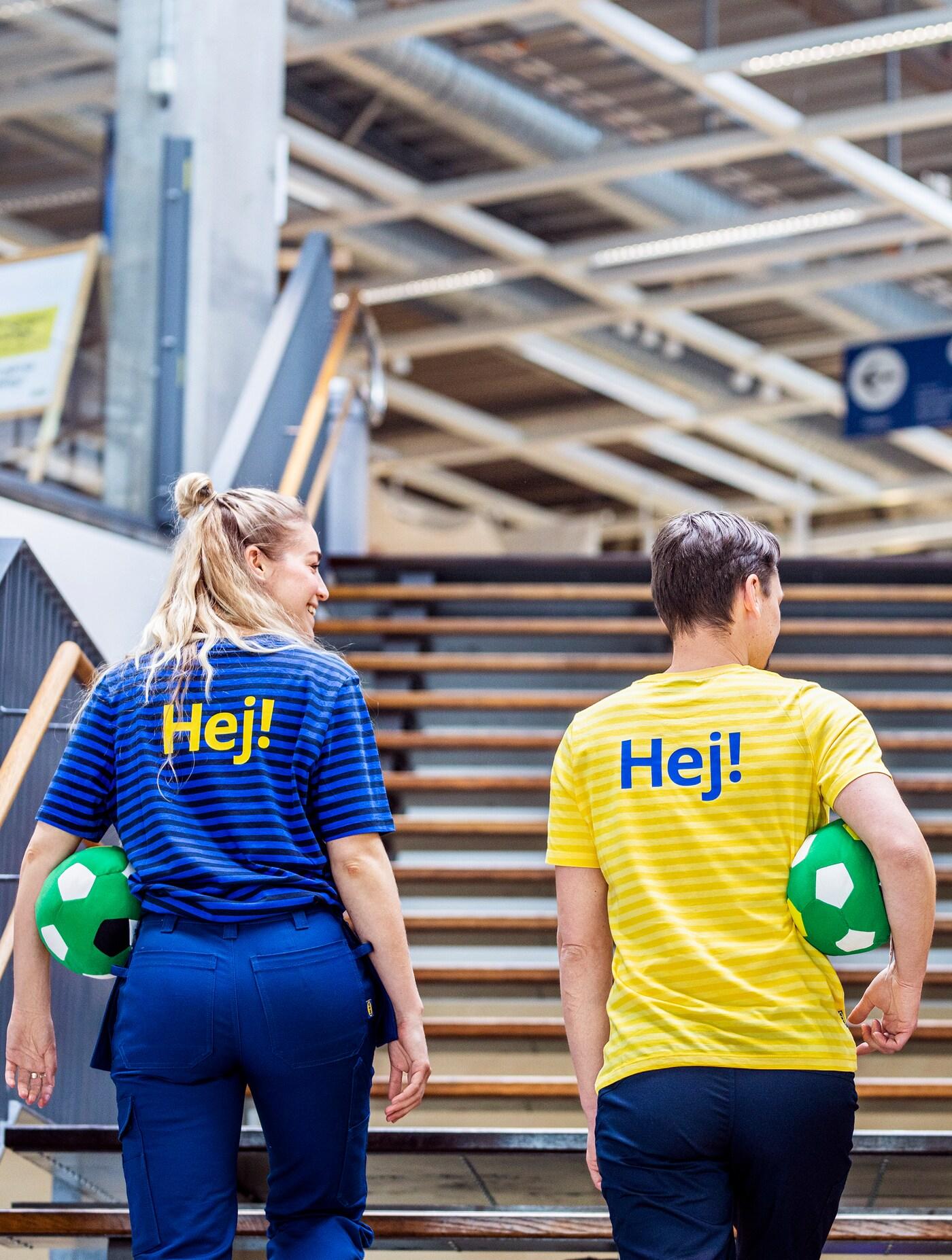 Two IKEA co-workers dressed in striped work clothes walk up wooden stairs with green footballs under their arms.