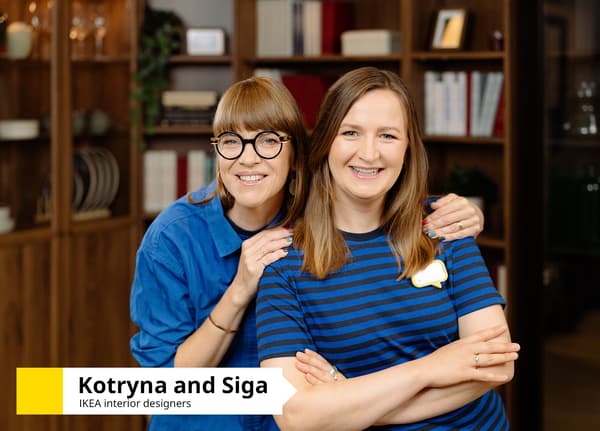Two IKEA interior designera wearing blue uniforms, standing in front of a dark wooden bookcase. Text in the bottom-left corner reads: “Kotryna and Siga, IKEA interior designers”.