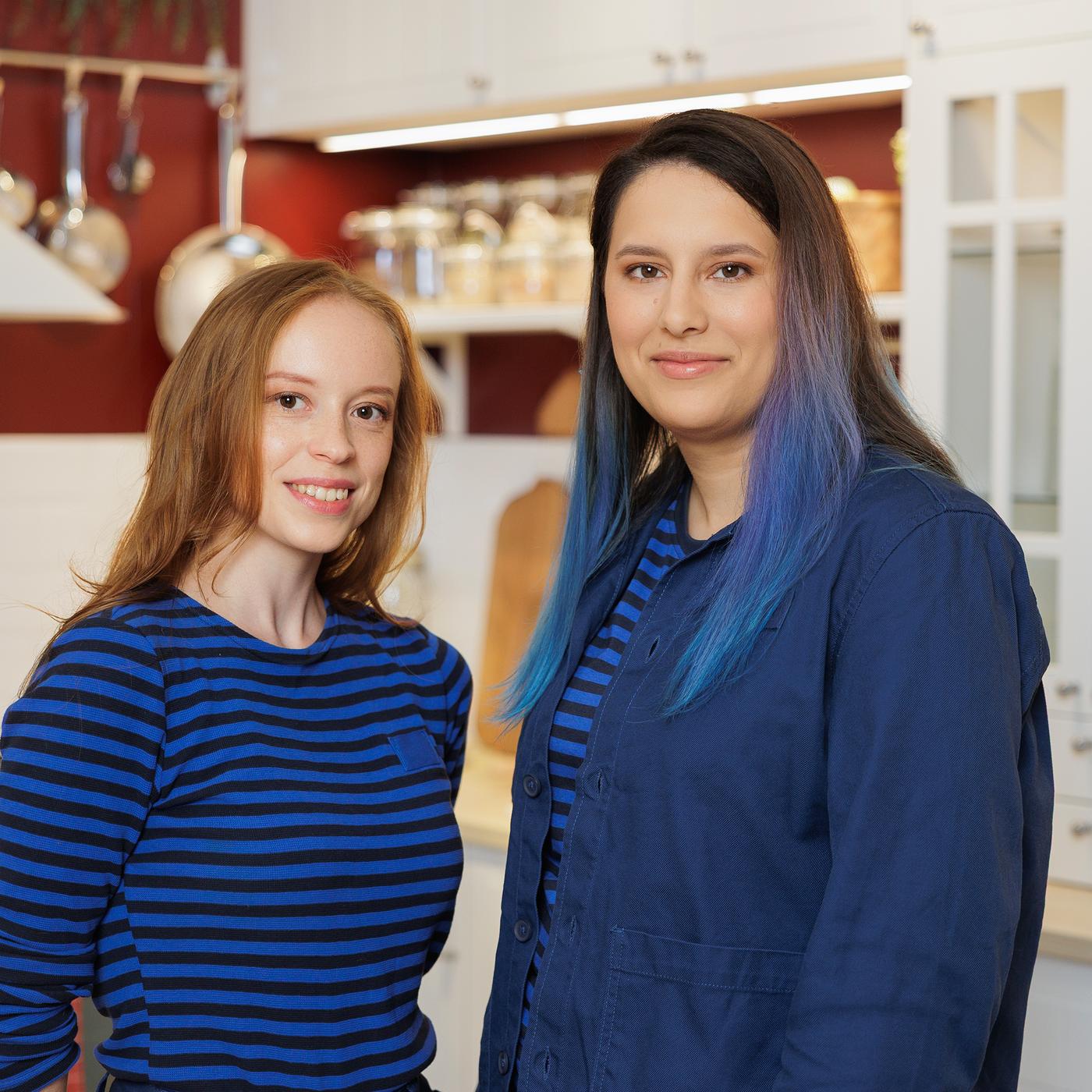 Two IKEA interior designers in blue striped uniforms stand confidently in a kitchen showroom with white cabinets and red walls.