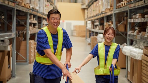 Two IKEA warehouse coworkers in striped shirts and safety vests moving a long flat pack between tall shelving in the storage area