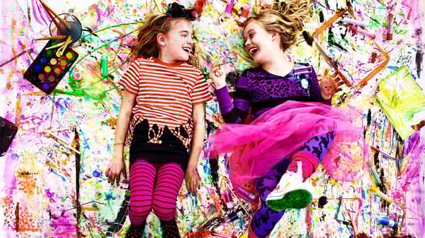 Two laughing girls lying on top of a giant paper with colour splashes and IKEA MÅLA watercolours beside them.