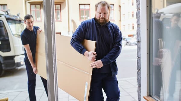 Two men carry a long, flat parcel through a front door.
