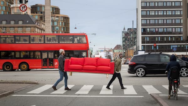 Two men carrying a red sofa across a pedestrian crossing in a city.