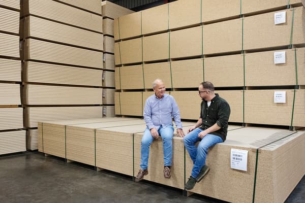Two men sit on a large pile of particle boards. All around them are particle boards stacked on top of each other.