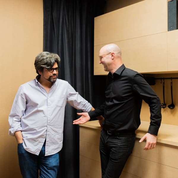 Two men stand talking in a beige room.