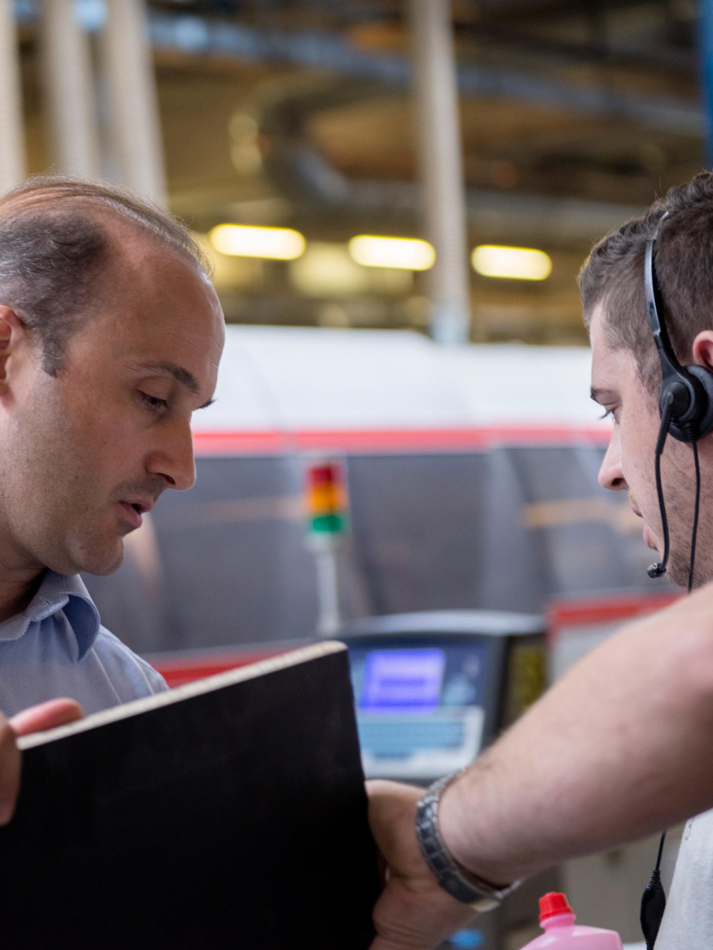 Two men standing beside some machinery in a factory, one carrying a lap top computer and the other wearing a headset.