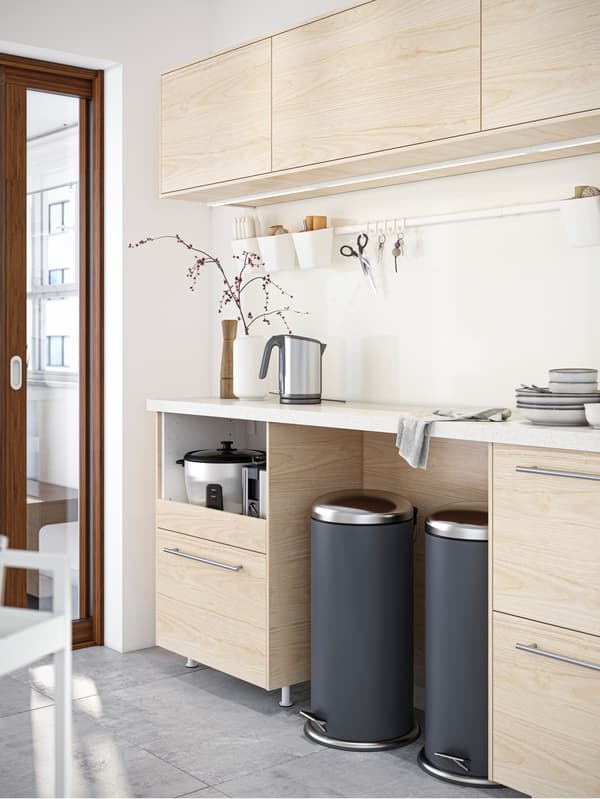 Two MJÖSA pedal bins stand under a kitchen worktop with a kettle and some tableware standing on it and cabinets above it.