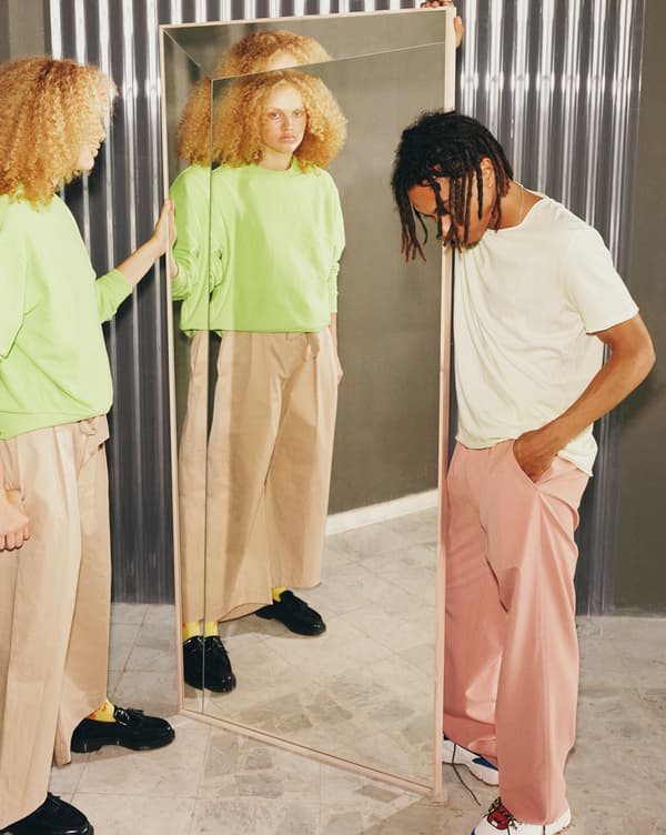 Two models hold a mirror with a long crack from the bottom to its top left corner, one of their faces refracted in it.