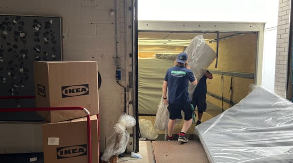 Two movers unload mattresses and boxes from a truck at a loading dock. The boxes are labeled IKEA.