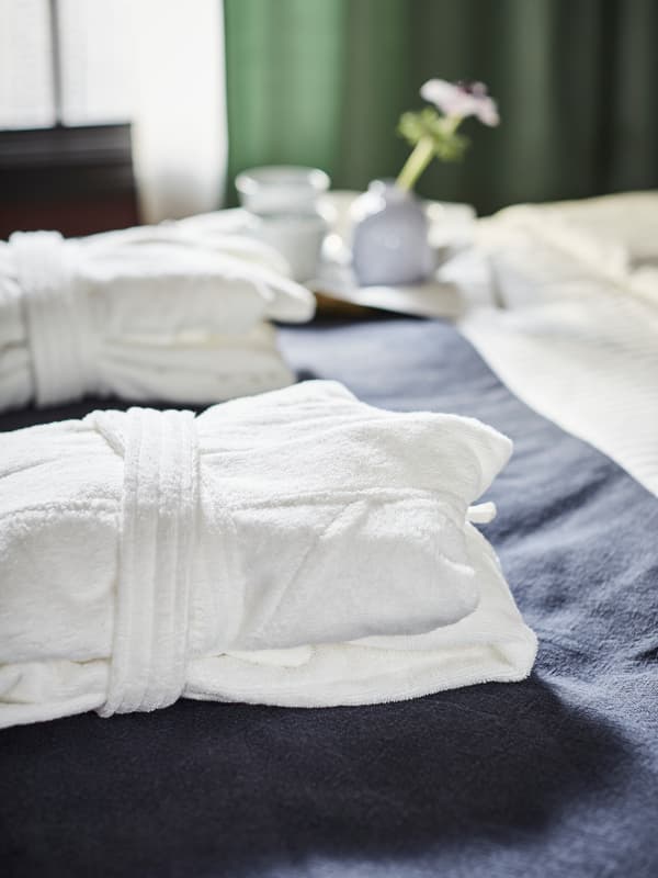 Two neatly folded white bathrobes on a made double bed; a coffee tray with two white cups, and a vase with a flower.