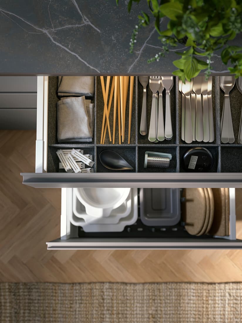 Two open kitchen drawers reveal neatly organized utensils and cutlery in the top drawer and stacked dishware in the bottom, with a green plant visible in the top right corner.