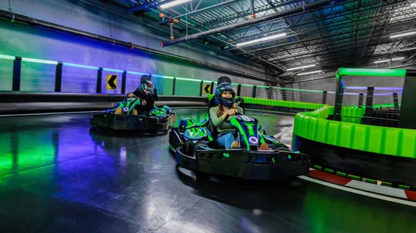 Two participants racing go-karts at Andretti Indoor Karting & Games with colorful track lights.
