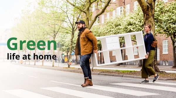 Two people are carrying a white KALLAX shelving unit threw the street. On picture it is written: Green life at home