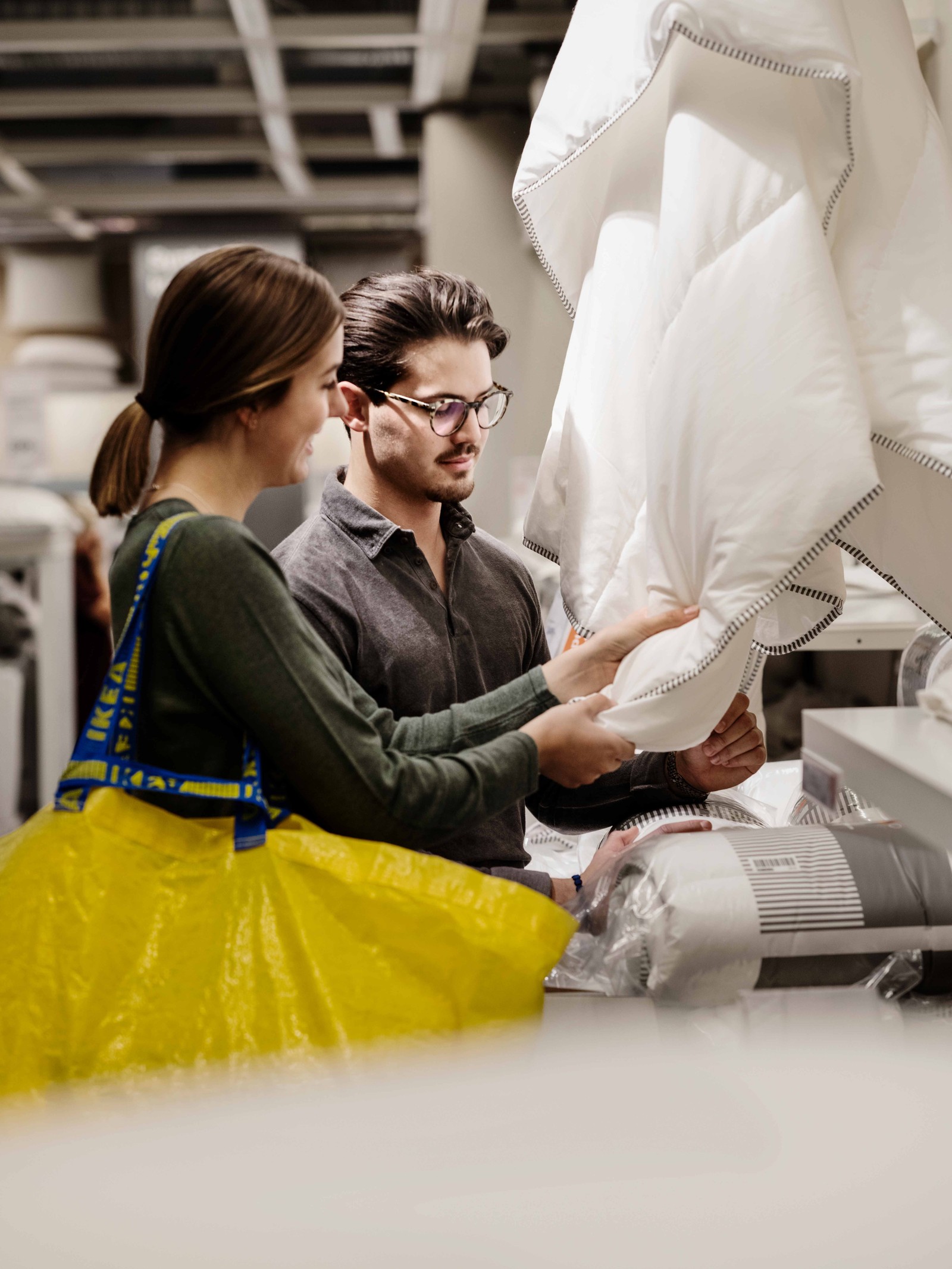 Two people are looking at blankets in the IKEA store and have a yellow IKEA bag with them.
