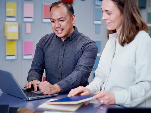 Two people are standing with their backs against a wall with different colourful patterns on it and looking at a laptop screen together