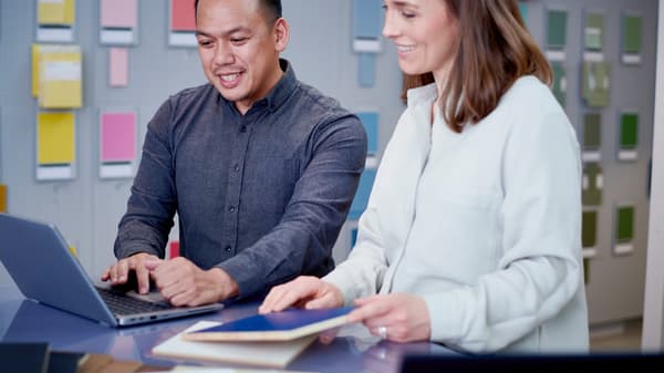 Two people are standing with their backs against a wall with different colourful patterns on it and looking at a laptop screen together.