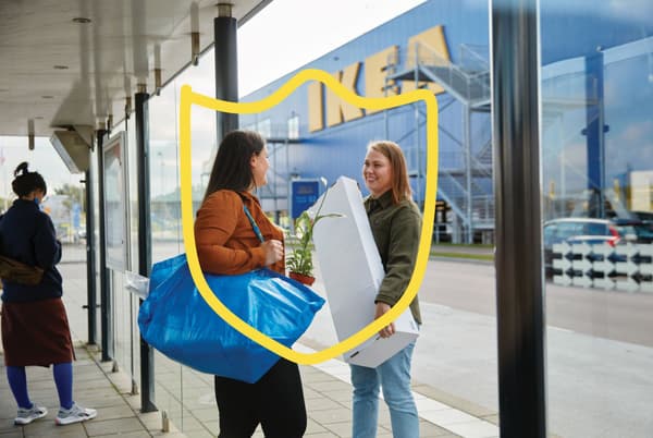 Two people at an bus stand holding items they have bought at the IKEA store across the street.