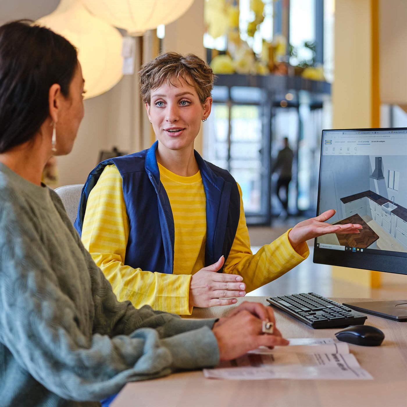 Two people discuss a 3D kitchen design displayed on a computer monitor in a modern office setting.